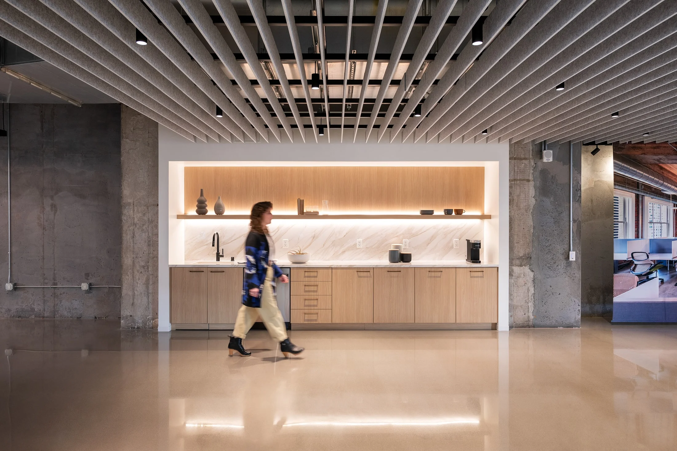 Office kitchenette by Quezada Architecture with linear wood cabinetry, integrated lighting, and an acoustic felt baffle ceiling.