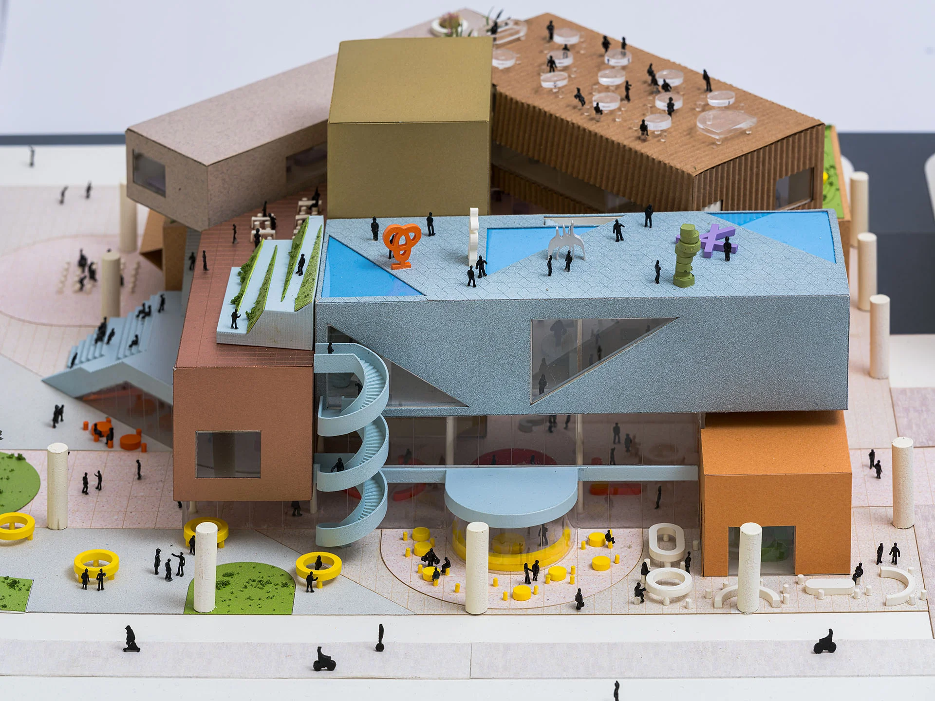 Physical architectural massing model by WORKac showing a multi-volume public complex with colorful roofs, spiral staircase, and site figures.