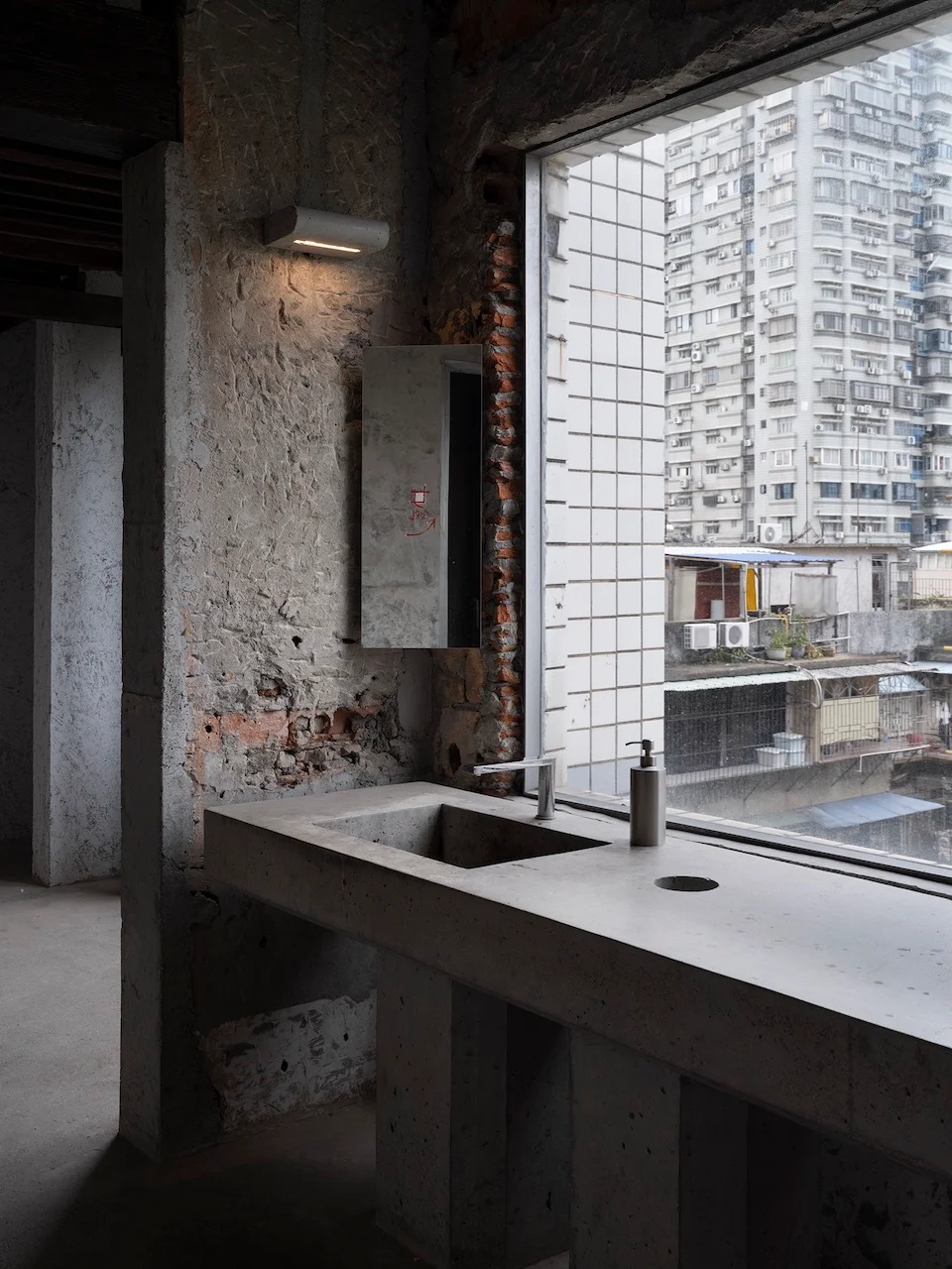 Concrete vanity and trough sink against a large window framing a dense urban view, with raw exposed masonry and plaster walls by devolution.