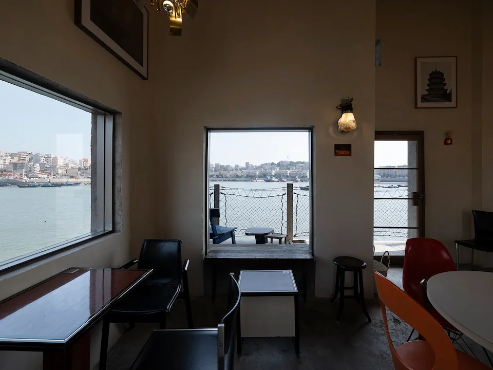 Interior of a waterfront cafe by 退化建筑devolution with framed views of the harbor through deep-set rectangular windows.