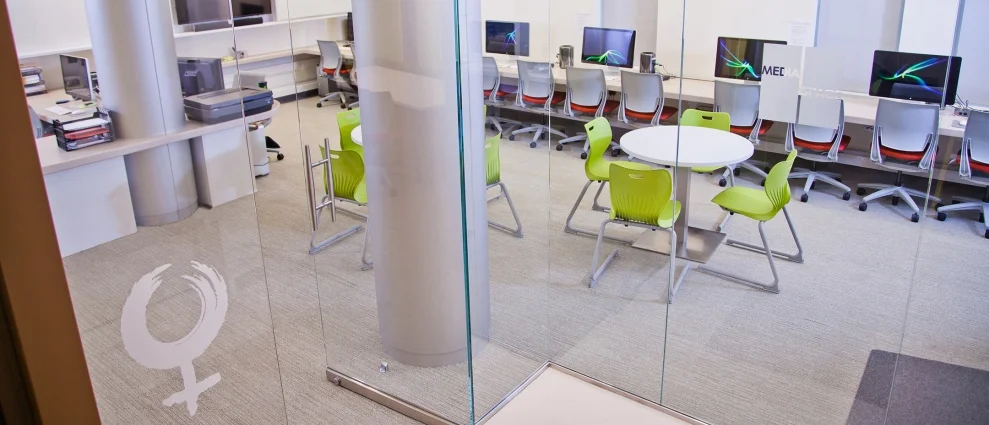 Interior view of an open workspace and media lab enclosed by floor-to-ceiling glass partitions, featuring computer stations and green chairs.