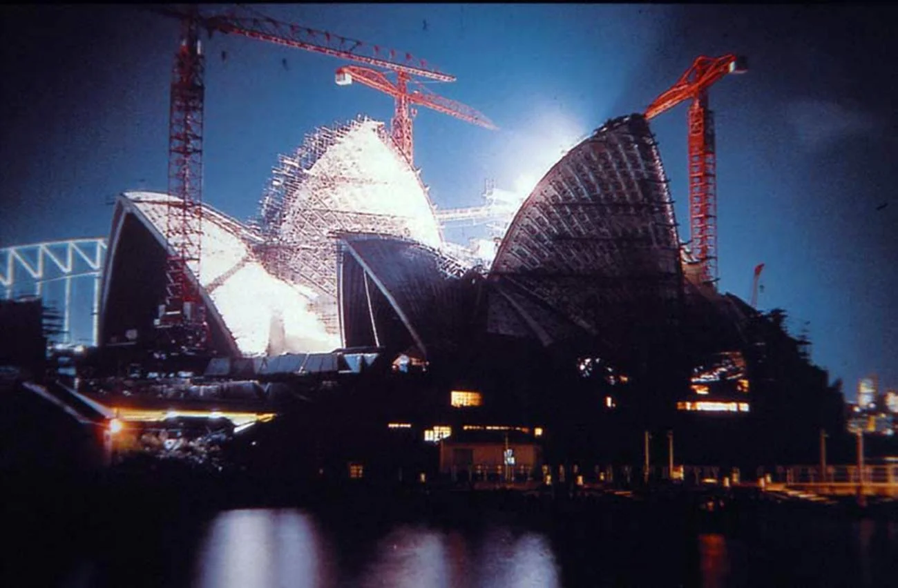 Night view of the Sydney Opera House under construction with illuminated concrete shells and cranes.