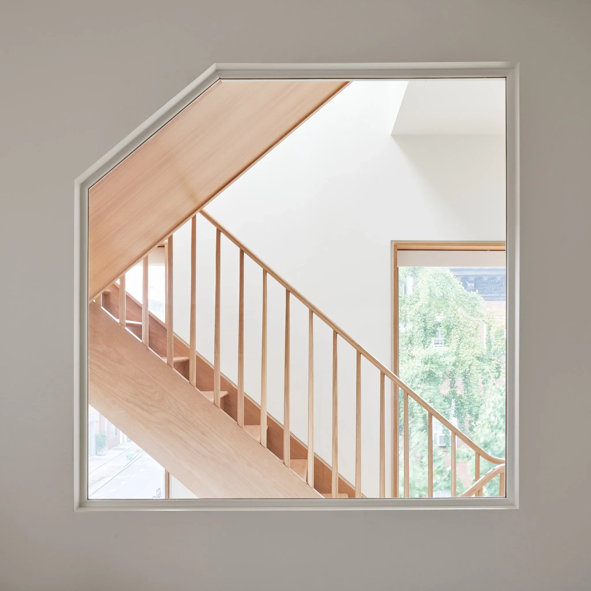Interior view through a faceted white framed wall opening revealing a wooden staircase by Sarah Jacoby Architect.