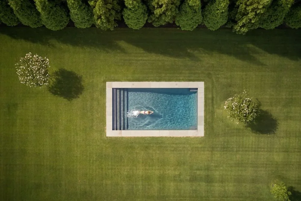 Overhead aerial view of a rectangular swimming pool embedded in a flat green lawn with trees by Sarah Jacoby Architect.