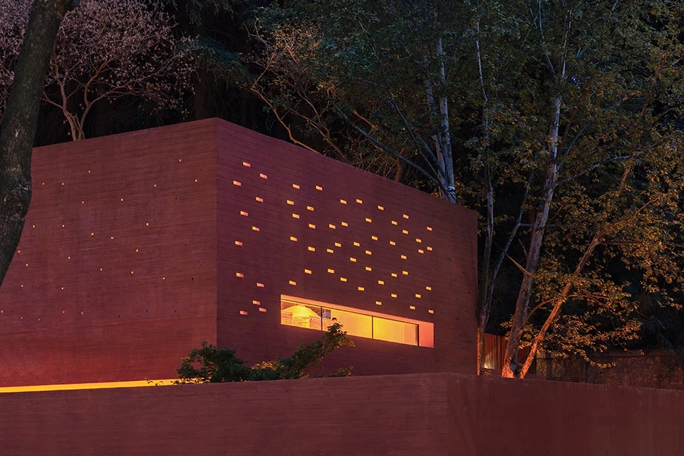 Night view of a reddish-brown board-formed concrete volume perforated with scattered small slot openings and a long horizontal window, by Mix Architecture.