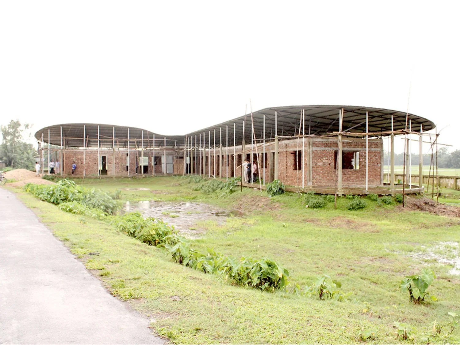 Low-slung brick community building with a sweeping double-curved roof supported by slender posts in a rural landscape.