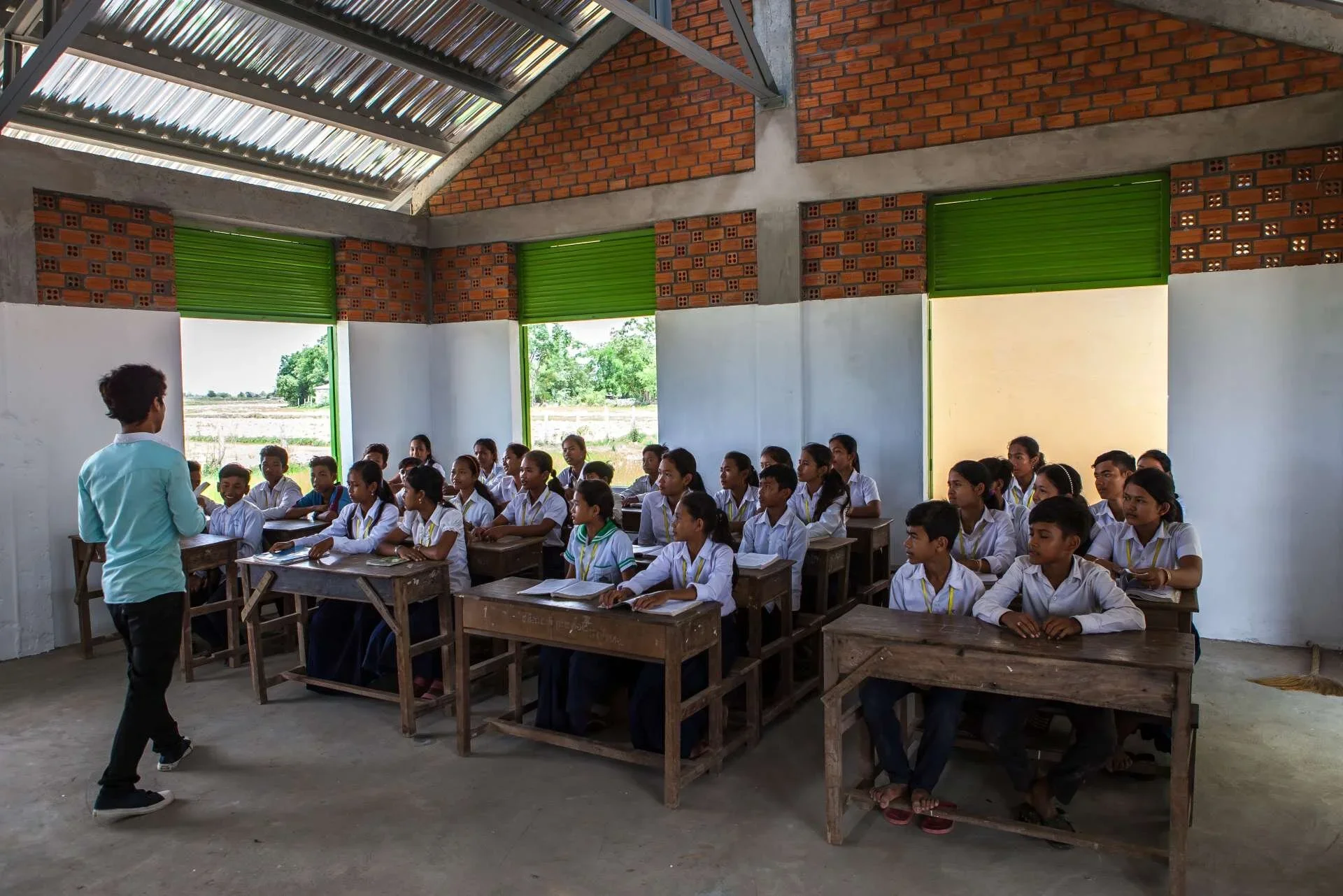 Interior of Khyaung School in Cambodia by Studio Jayga Architects showing a classroom with students, exposed brick walls, and corrugated metal roof.