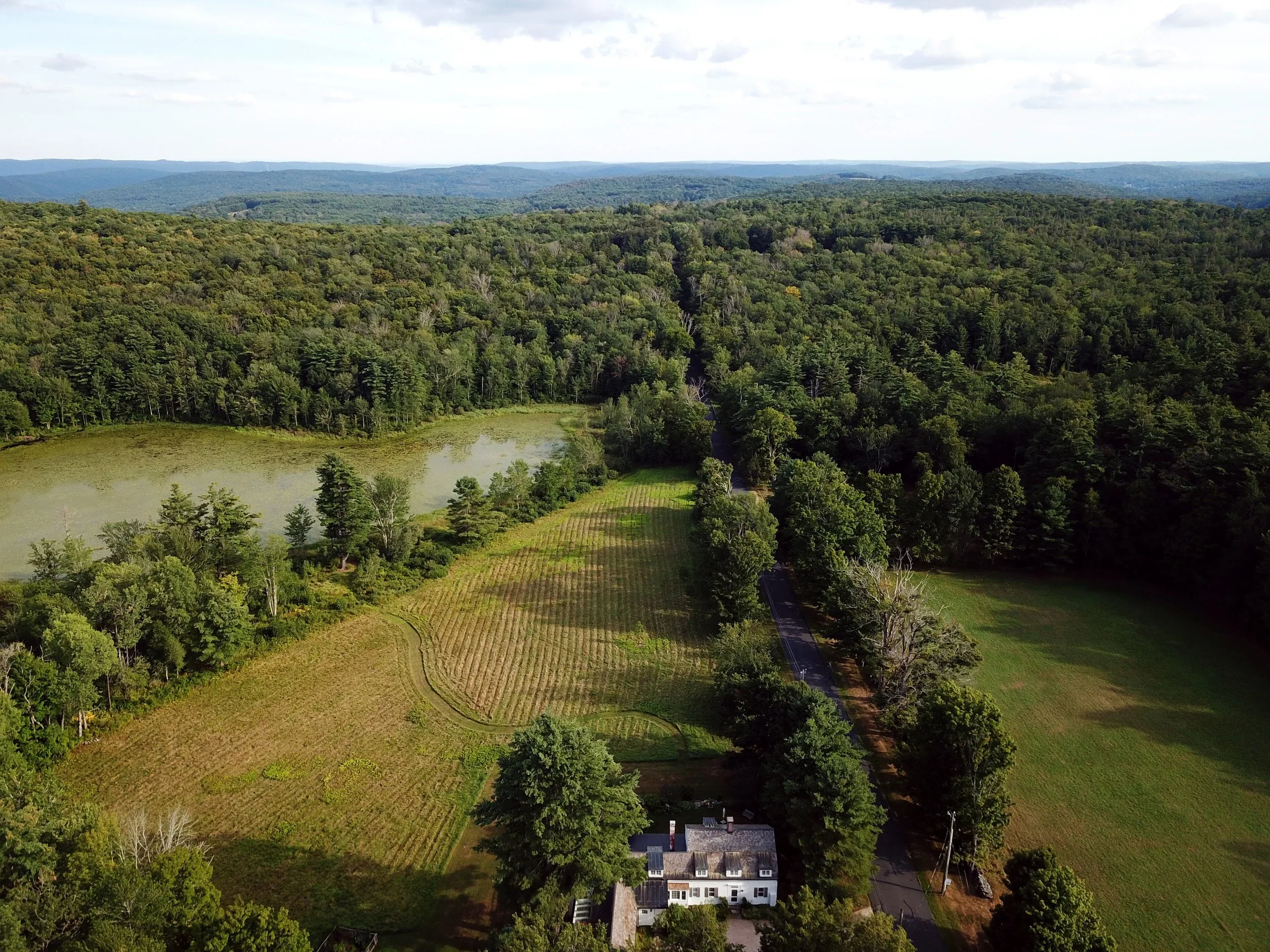Aerial drone view of a rural landscape with a farmhouse, fields, pond, and dense forest by Path Place Architecture.