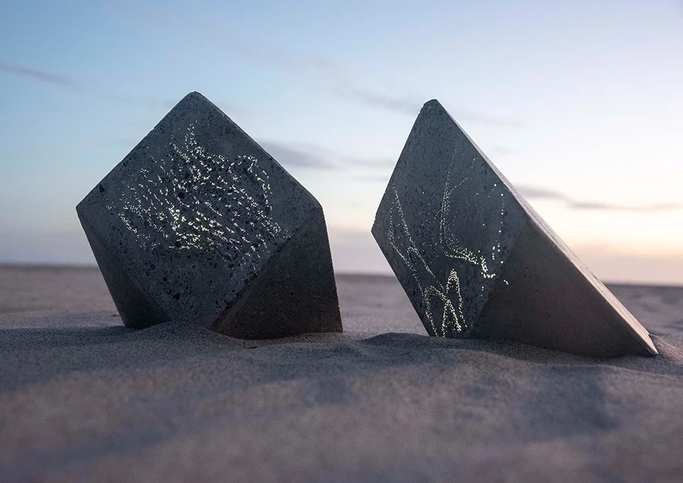 Two slanted concrete cubic lighting fixtures with glowing fiber-optic constellations embedded in their faces, set in sandy ground at dusk by Epiphany Architects.