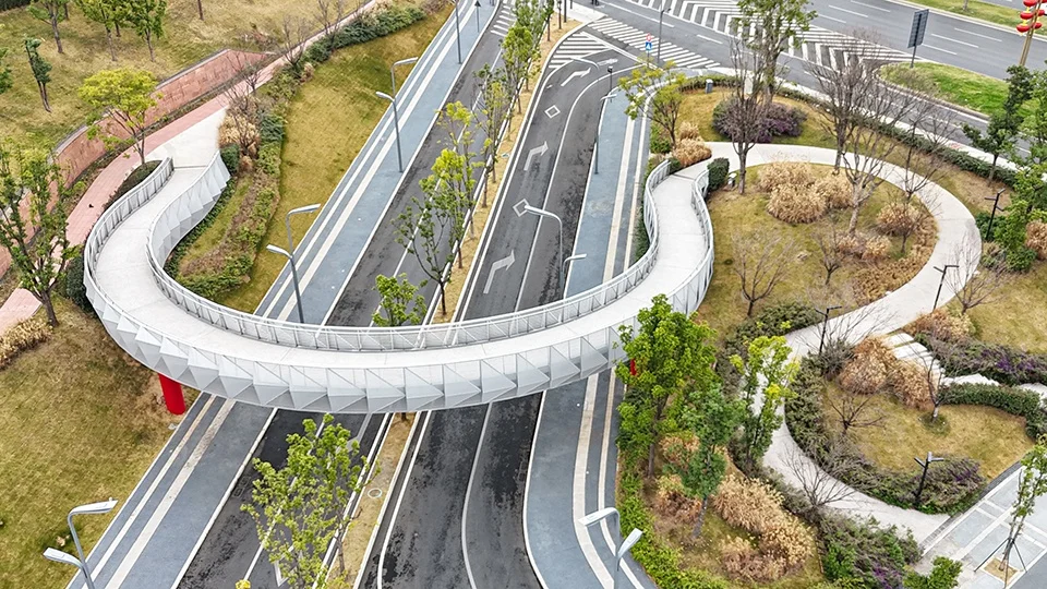High-angle aerial view of a curving pedestrian overpass with patterned metal cladding spanning across a multi-lane roadway.