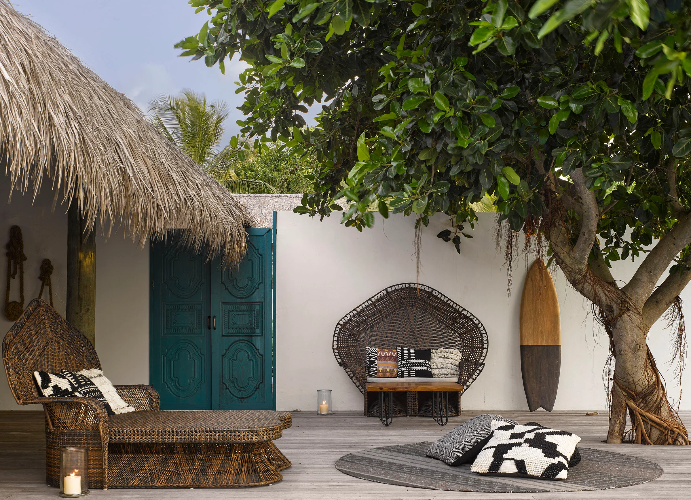 Outdoor tropical resort lounge terrace with thatched roof, teal double doors, woven furniture, and a large tree by Studio Carter.
