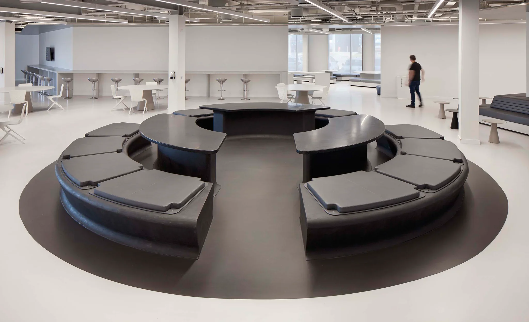 Large open plan office interior by PENSON featuring a central circular curved black communal seating and table installation.
