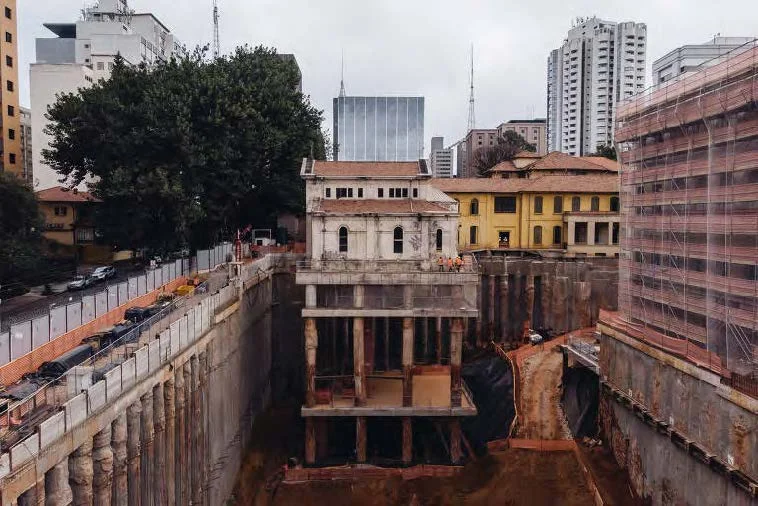 Aerial view of a historic building hoisted on tall concrete piers above a massive deep excavation pit in an urban setting by Triptyque.