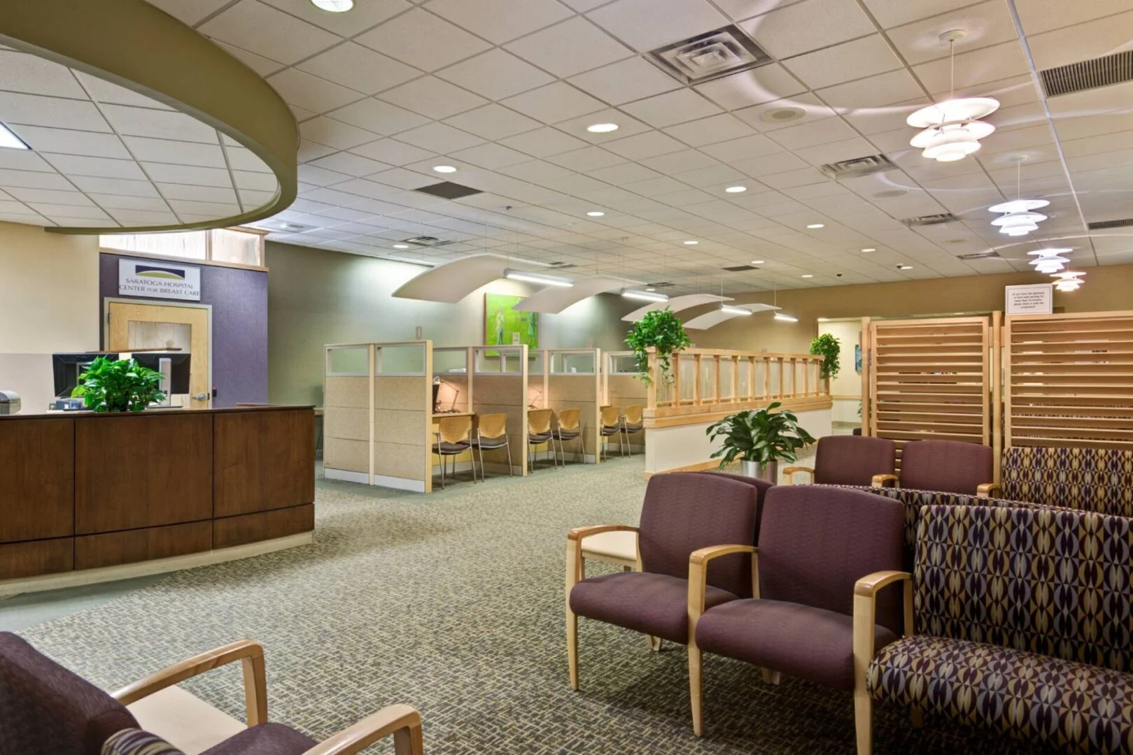 Waiting and reception area of a healthcare clinic with carpeted flooring, acoustic ceiling tiles, seating, and semi-private workstations.