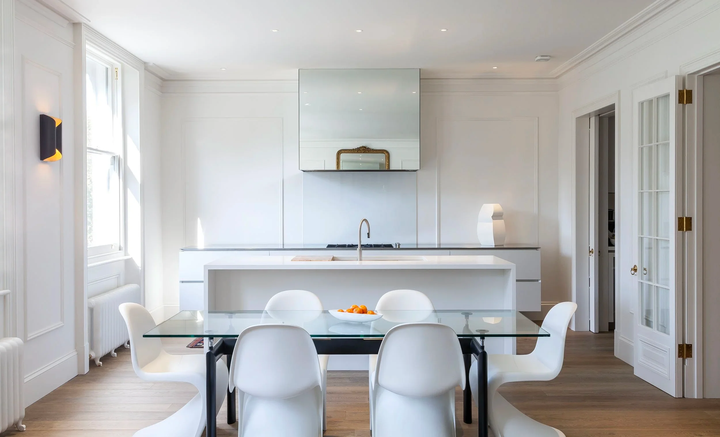 White minimalist kitchen and dining area with glass table and white molded chairs in a Queens gate apartment by Bolton Chalklin Architects.