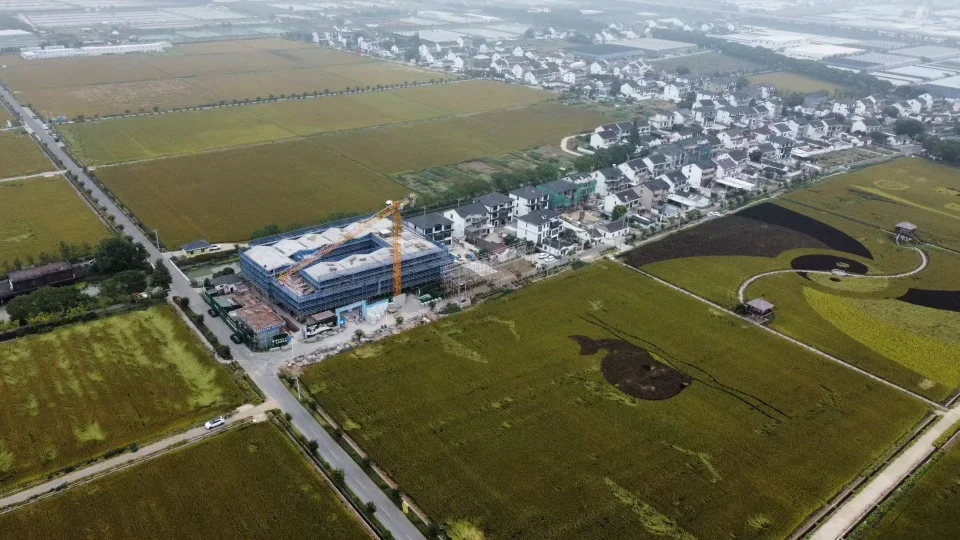 Aerial view of a rural construction site surrounded by agricultural fields and low-rise village houses, with a crane on site.