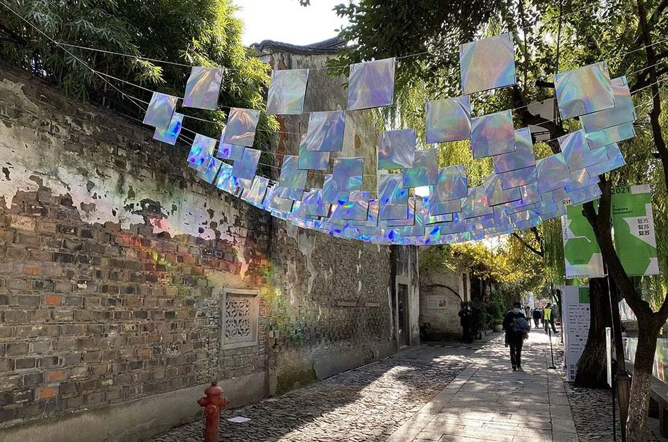 An outdoor urban alleyway featuring an overhead catenary installation of shimmering iridescent panels casting rainbows on a weathered brick wall.