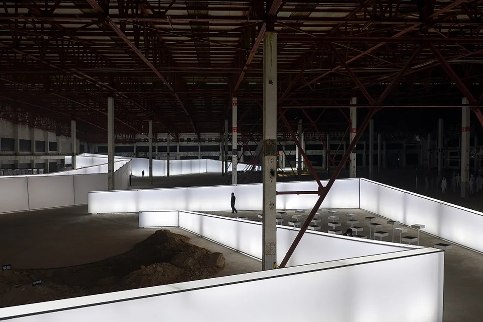 Wide interior shot of Atelier Xi exhibition design featuring glowing serpentine light walls inside a large industrial shed.