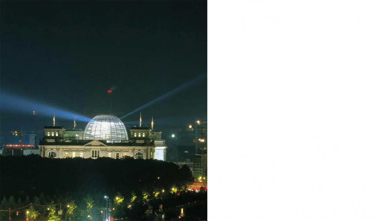 Night view of the Reichstag building in Berlin with its glowing glass dome and projecting light beams.