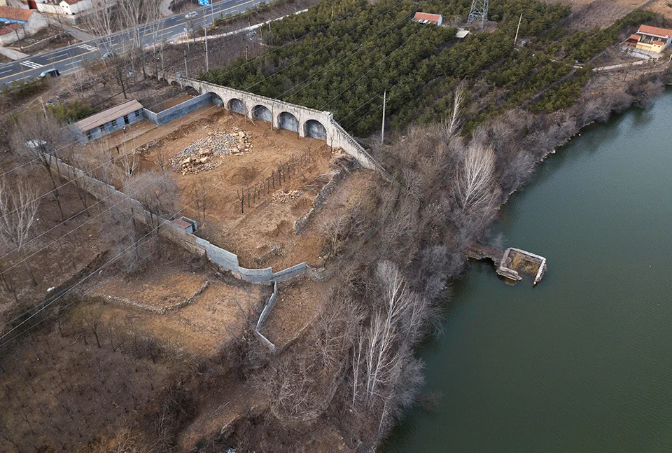 Aerial view of a water reservoir site with an old masonry arched structure and bare trees by xiān氙建筑工作室.