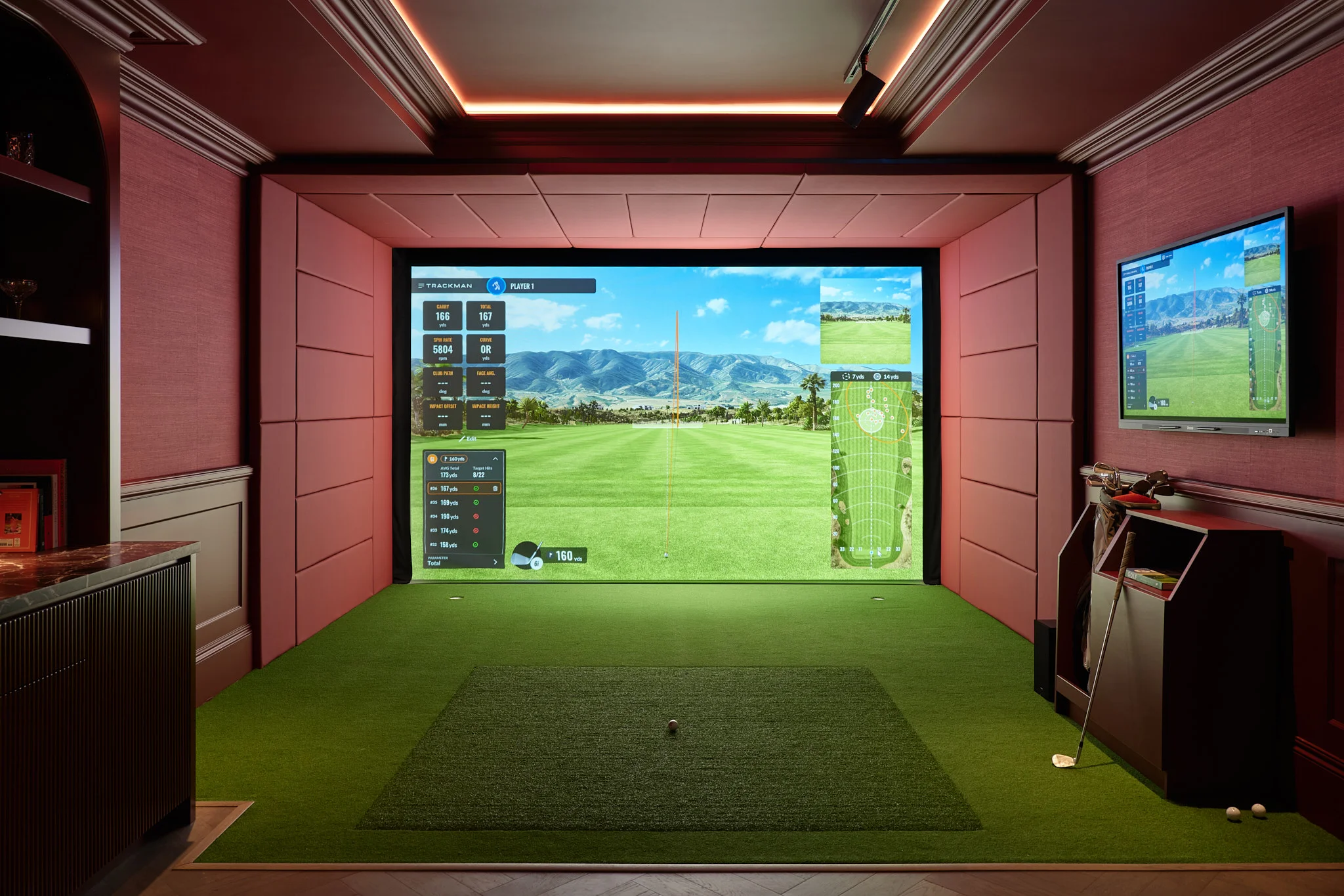Interior shot of a luxury golf simulator room with deep red fabric walls, a large projection screen, and a green turf floor by SGS Design.