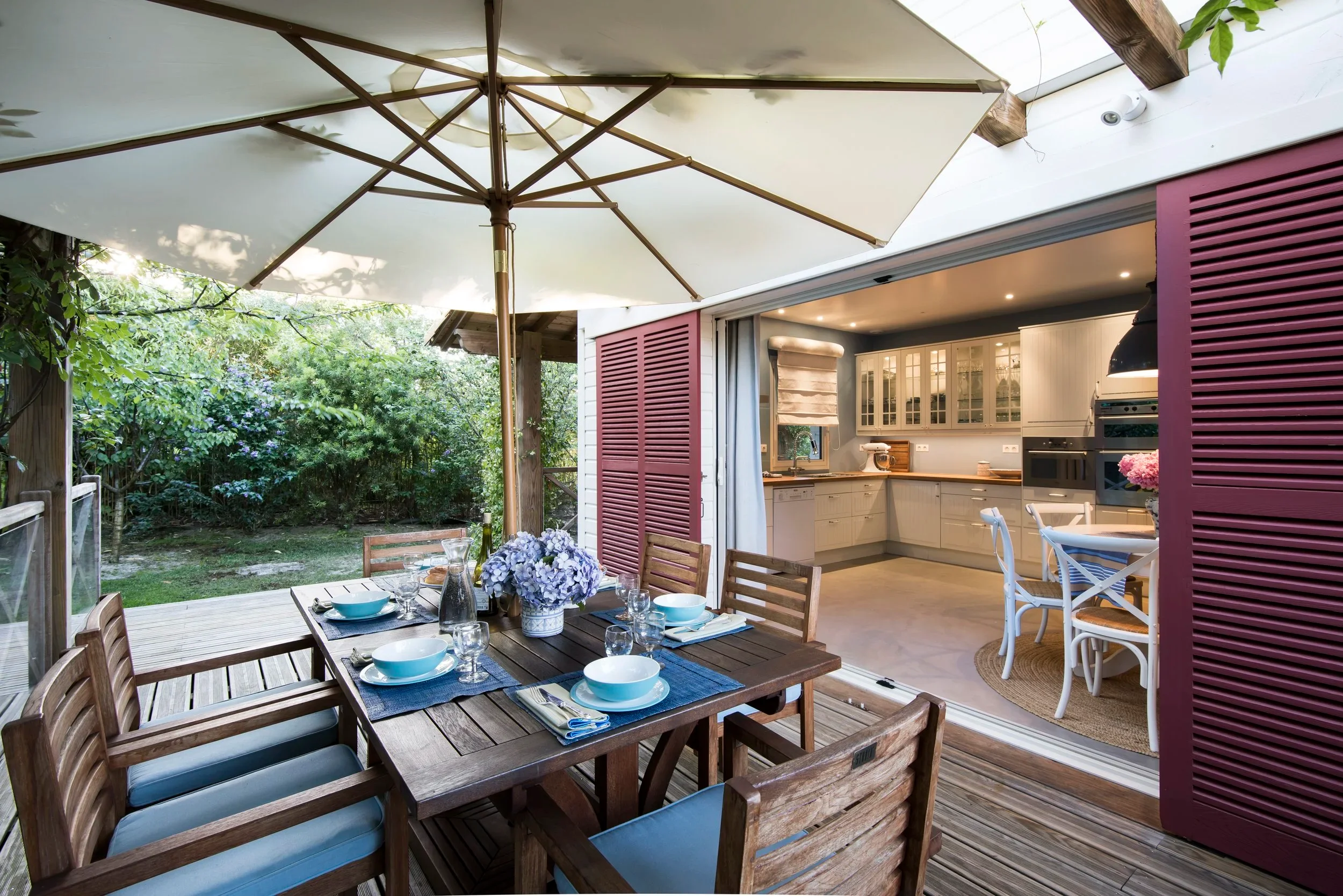 Covered outdoor dining terrace with large umbrella opening into a kitchen interior with red louvered shutters by Claudia Dorsch Interior Design.