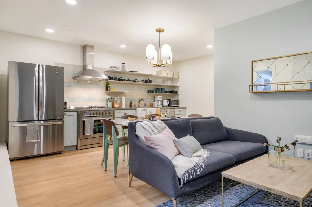 Open-plan living room and kitchen interior with pale blue cabinetry, stainless steel appliances, and a dark blue sofa by OpenScope Studio.