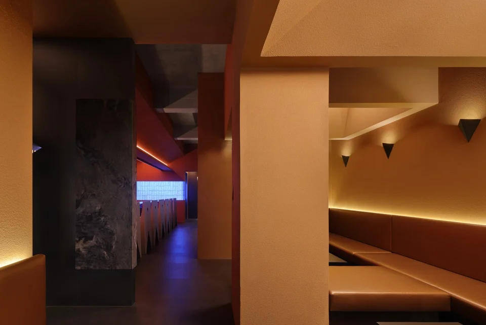 Interior view of Origami bar in Shanghai by Tens Atelier, featuring warm amber banquette seating, textured walls, and contrasting blue-lit corridor elements.