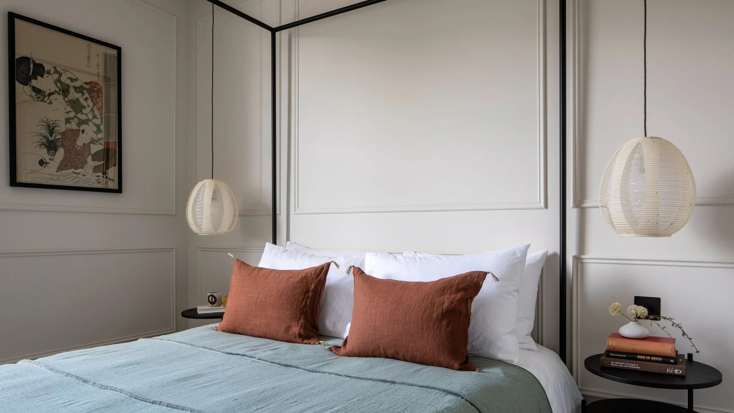 A calm bedroom interior featuring a black metal canopy bed frame, white walls with classical wall paneling, and sage and terracotta textiles by Katie McCrum Studio.
