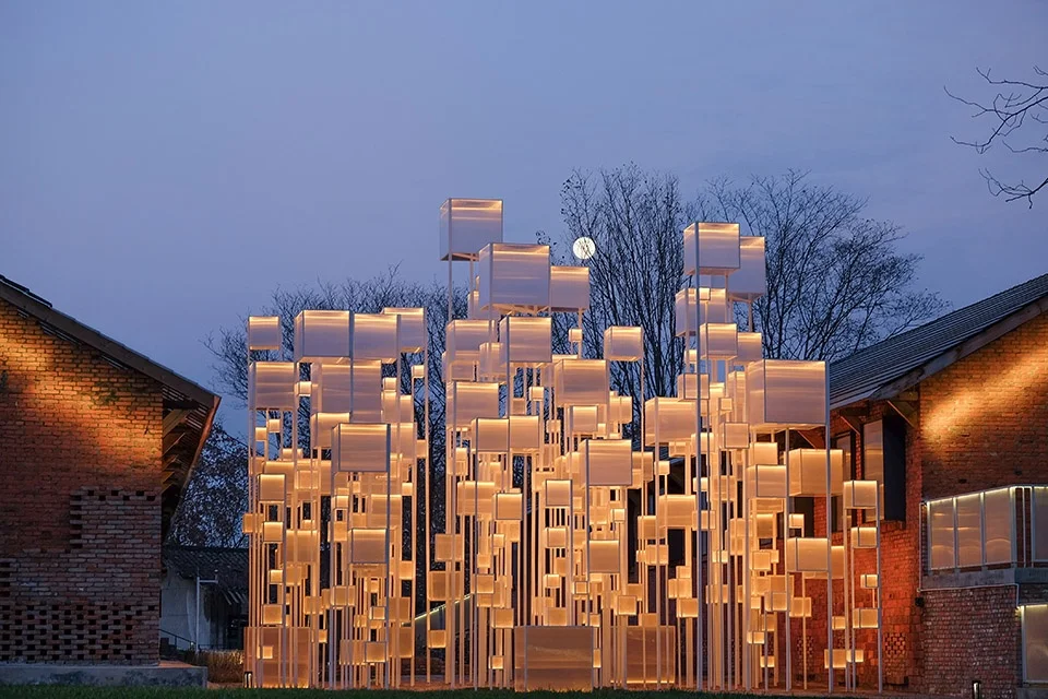 An outdoor installation by Archermit of glowing, staggered cubic volumes on slender posts between two historic brick buildings at twilight.