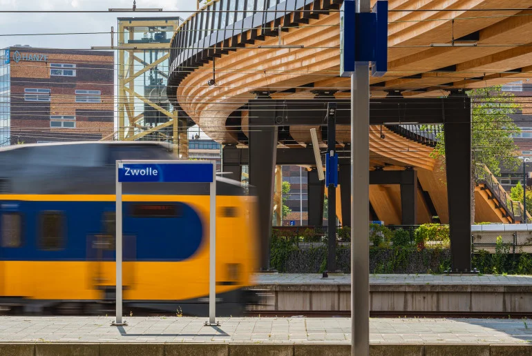 Elevated curving timber bicycle bridge structure with steel columns above a railway station platform with a passing train.