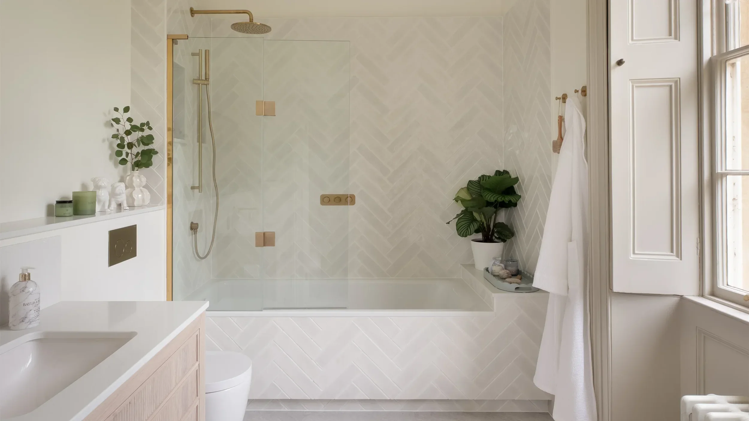 Bright bathroom interior with white herringbone tiles around a bathtub-shower combo, brushed brass fixtures, and a wooden vanity by Hobson's Choice.