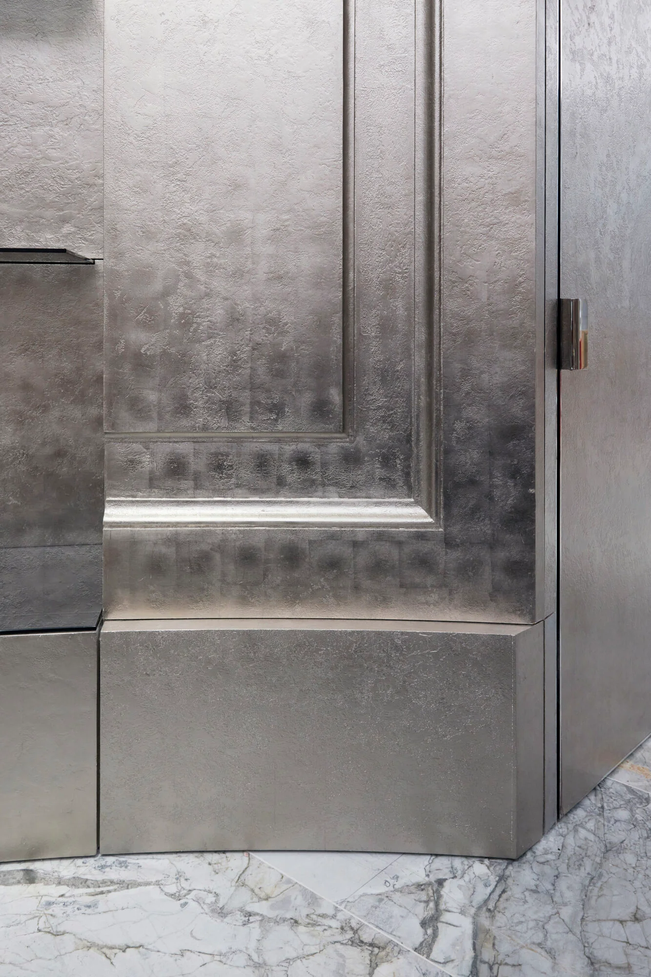 Close-up detail of textured metallic silver-leaf wall paneling and a flush door with a slim metal handle, set above a white marble floor by Casper Mueller Kneer.
