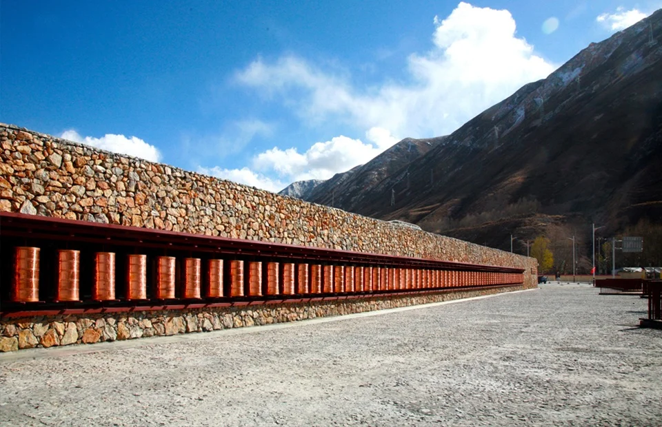Long stone memorial wall with a continuous line of suspended copper prayer cylinders under a bright sky and mountain backdrop.