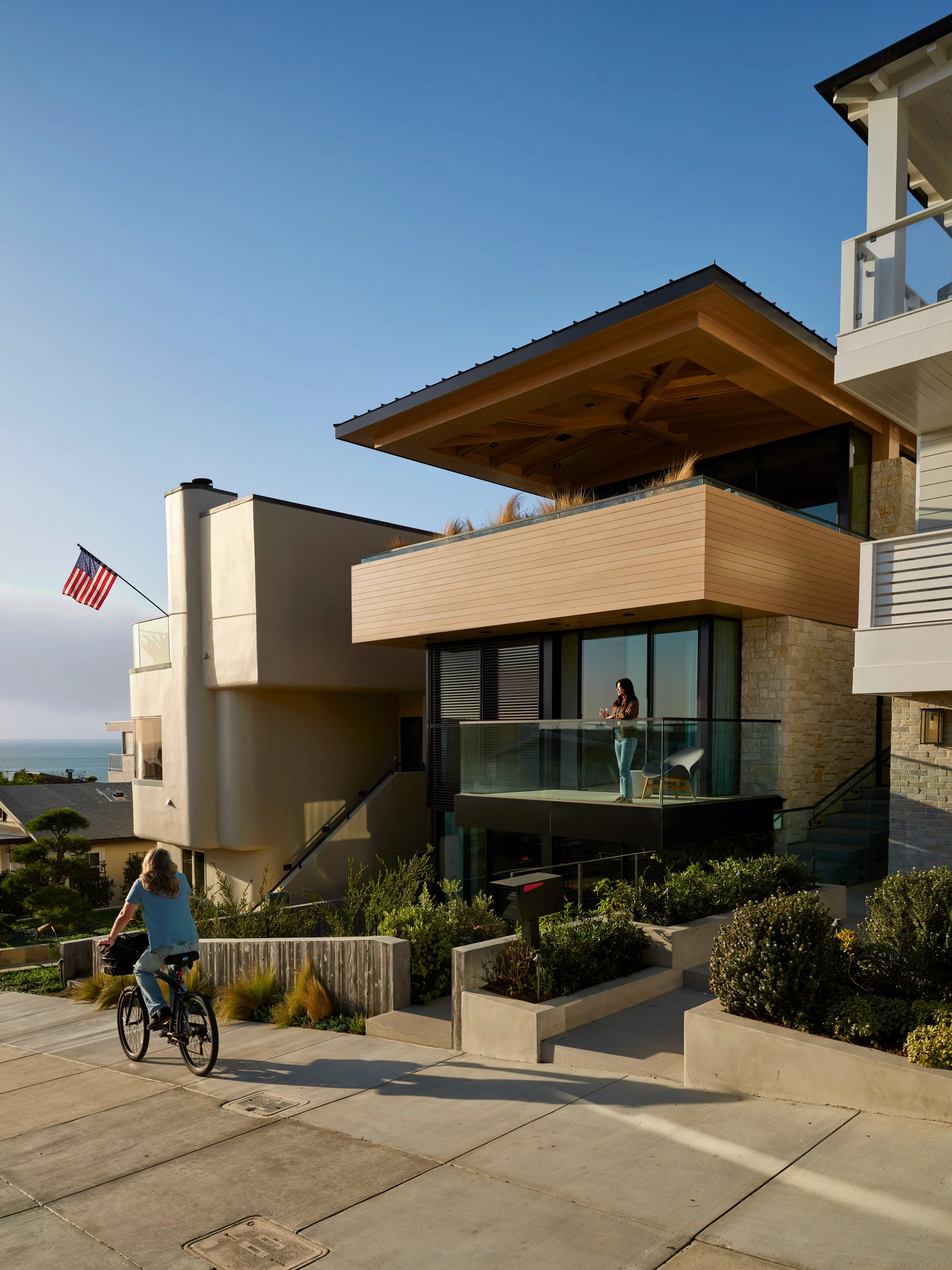 Street-facing exterior of HA Residence by Laney LA featuring cantilevered timber roof, stacked stone, glass balconies, and coastal landscape.
