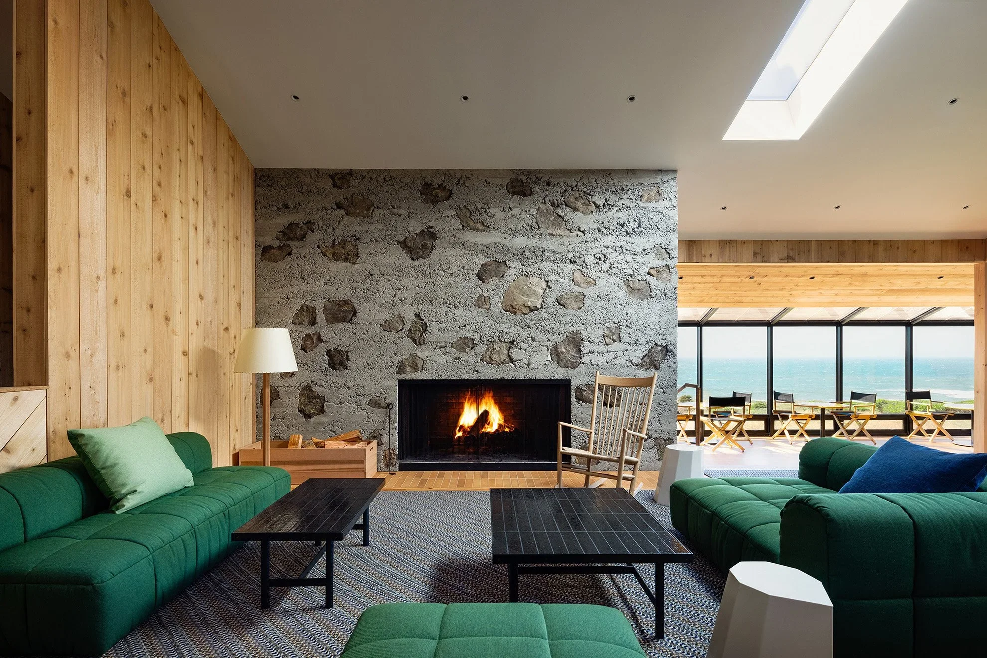 Lodge interior living room with a rough stone fireplace, vertical wood paneling, and green modular sofas looking out toward the ocean at Sea Ranch Lodge by Charles de Lisle.