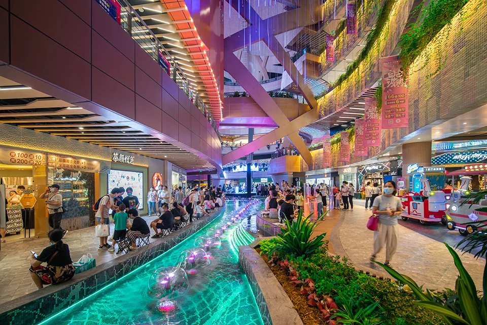 Interior atrium of a shopping mall featuring a winding water feature with neon lighting, multi-level balconies, and angled structural columns by COLORFULL.