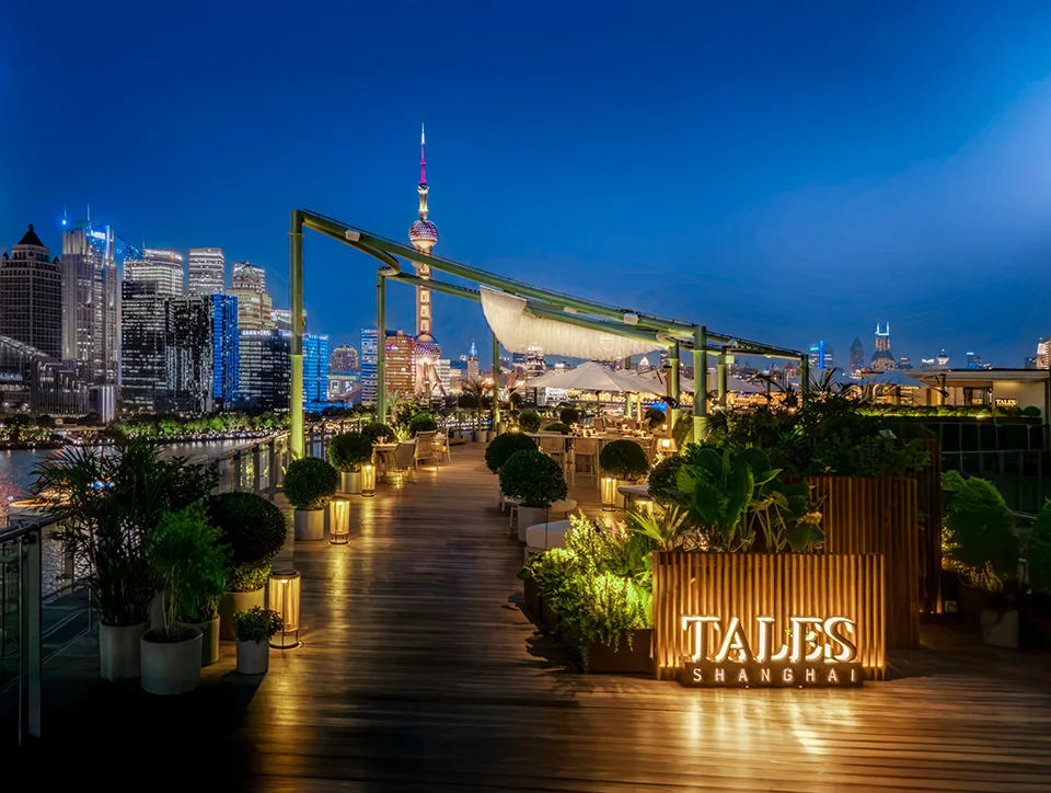 Rooftop terrace bar with wooden decking, lush planters, and glowing signage overlooking the Shanghai skyline at twilight.