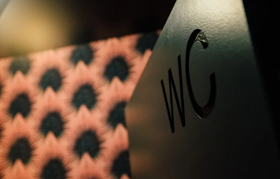 Close-up detail of a dark metal restroom sign with cut-out WC lettering against patterned wallpaper.