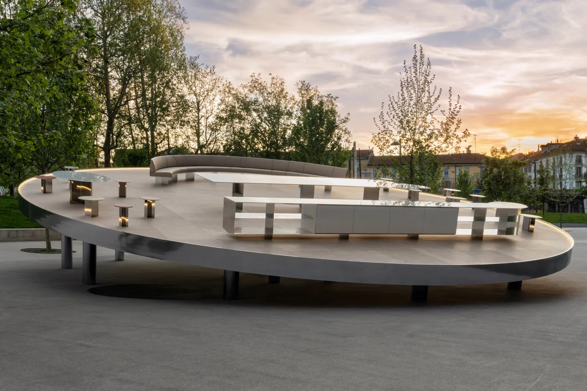 An elevated elliptical outdoor pavilion with integrated white tables, curved seating, and glowing base elements by Studio Klass.