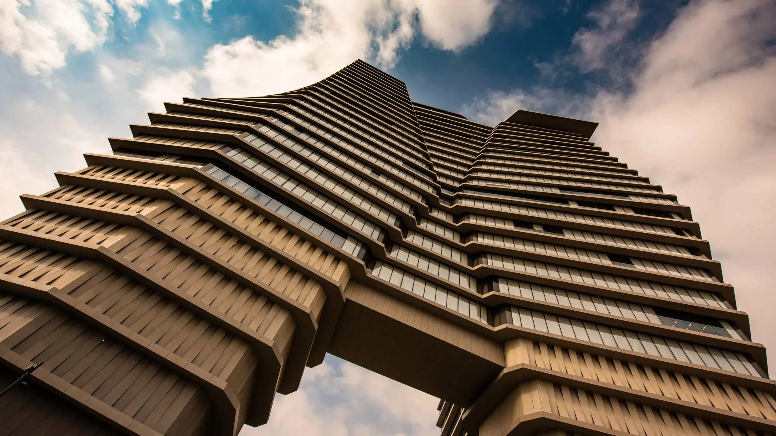 Worm's-eye view of a monumental skyscraper with stepped, cantilevered concrete floor plates and a central skybridge by Ron Arad Studio.
