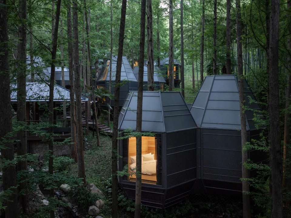 Dark metal faceted cabin modules nestled within a dense forest by GLA Architecture, showing a warm glowing bedroom interior.
