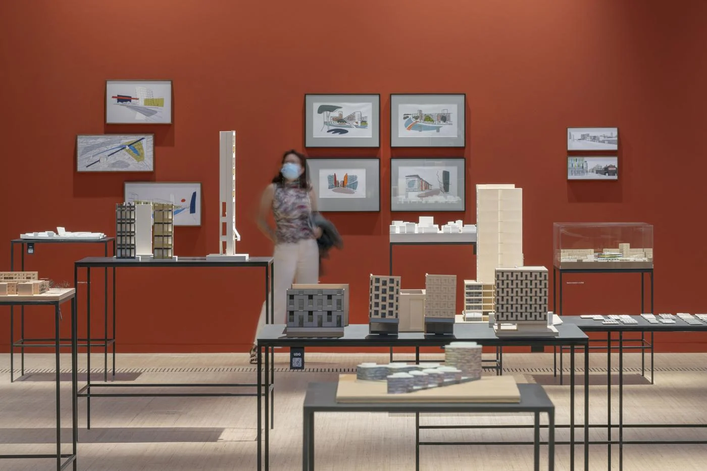 An exhibition interior displaying architectural models and drawings on slender dark metal tables against a terracotta-red wall by Sauerbruch Hutton.
