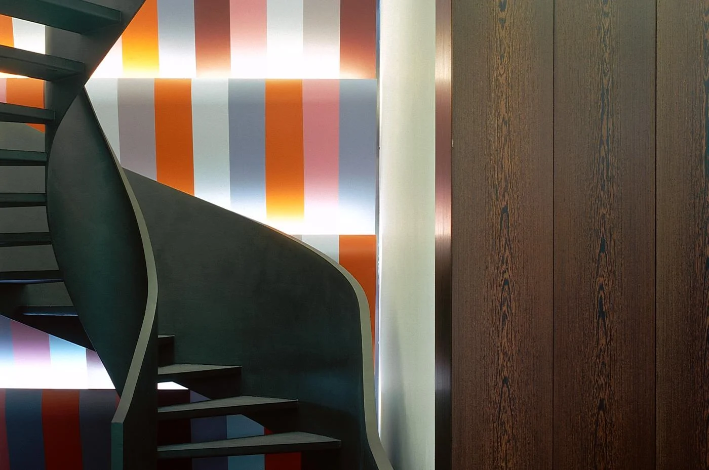 A dark curved metal spiral staircase set against a backlit vertical multicolored striped wall, beside dark wood paneling by Sauerbruch Hutton.