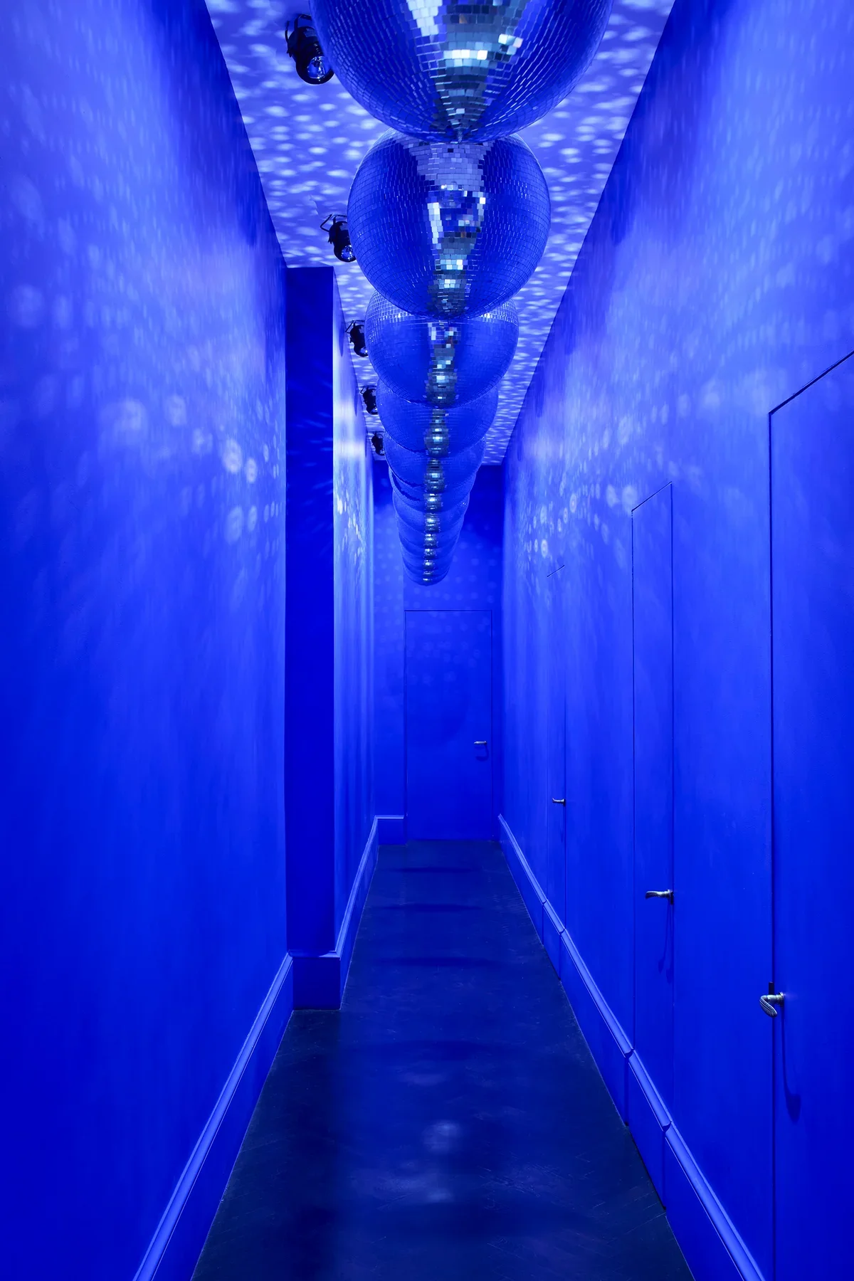 Monochromatic blue hallway lined with floor-to-ceiling doors and a central column of hanging disco balls reflecting light onto walls and ceiling.